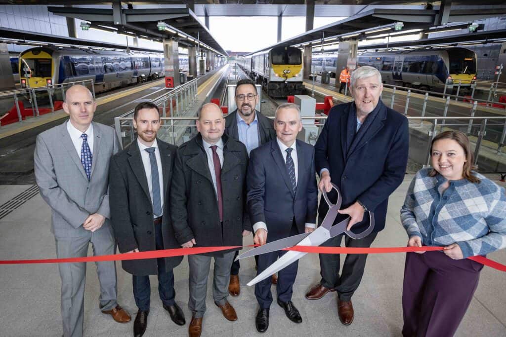 Group of smiling Babcock, Translink and NI Assembly representatives with a train platform behind them cut a red ribbon with large paper scissors