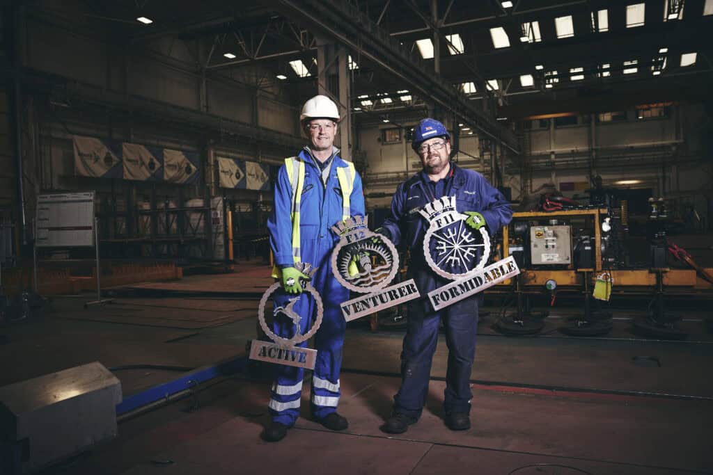 Two Babcock employees in blue overalls holding the crests for each of the three Type 31 frigates - Active, Venturer and Formidable