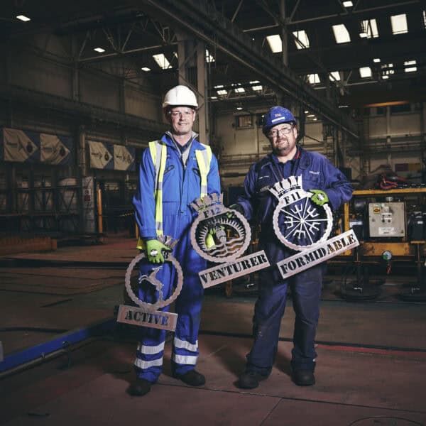 Two Babcock employees in blue overalls holding the crests for each of the three Type 31 frigates - Active, Venturer and Formidable